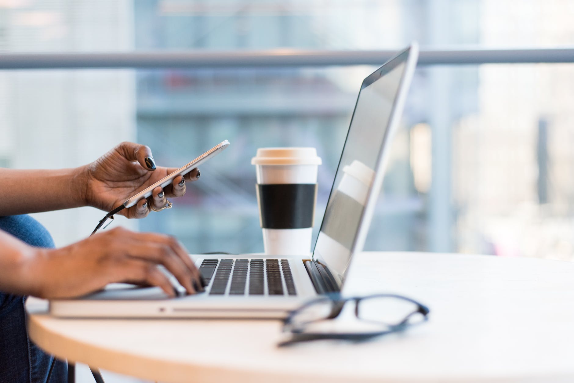 A female typing on a laptop while checking her smartphone during coffee time.