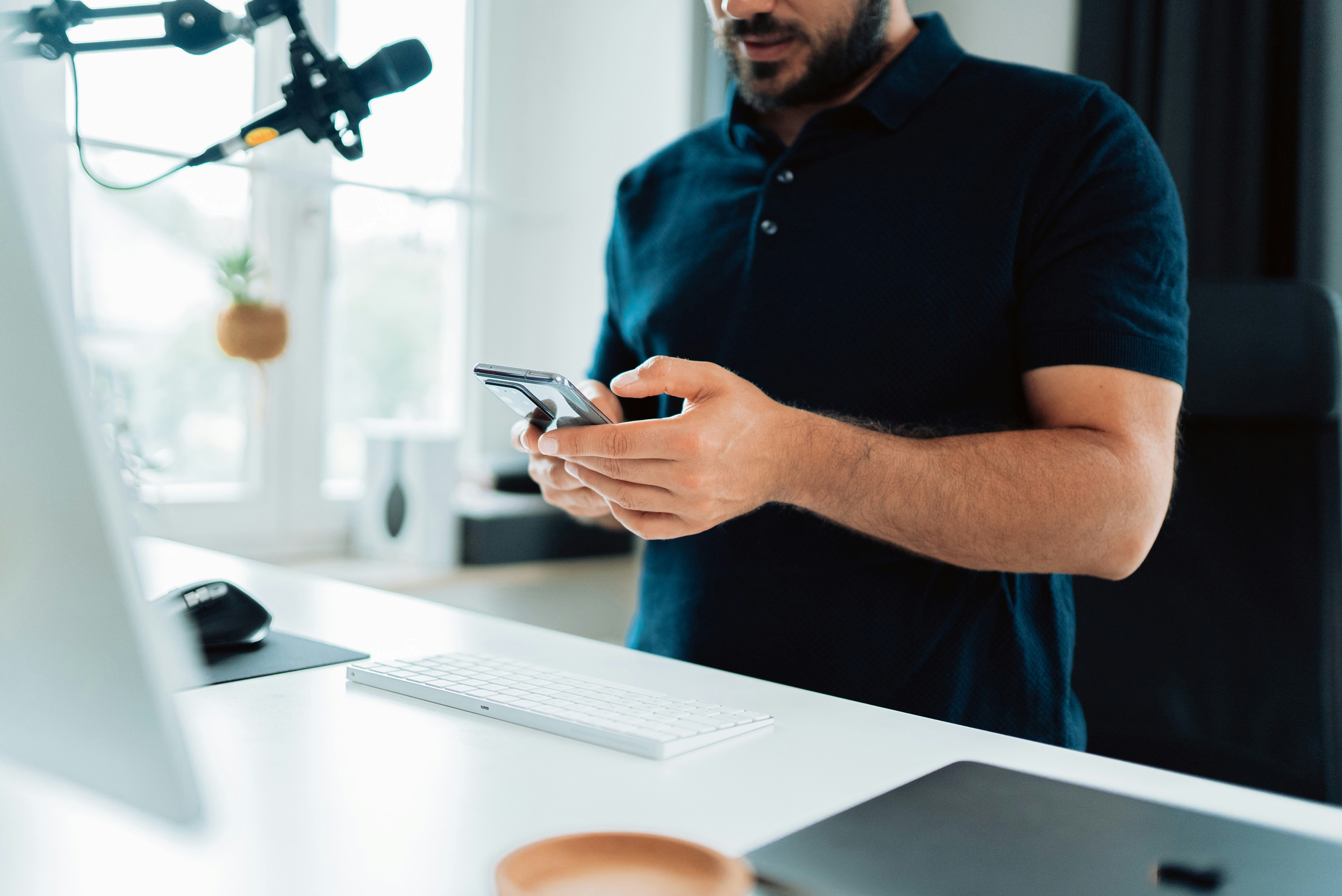 Man in blue polo shirt holding silver iPhone 6 over his white working desk.